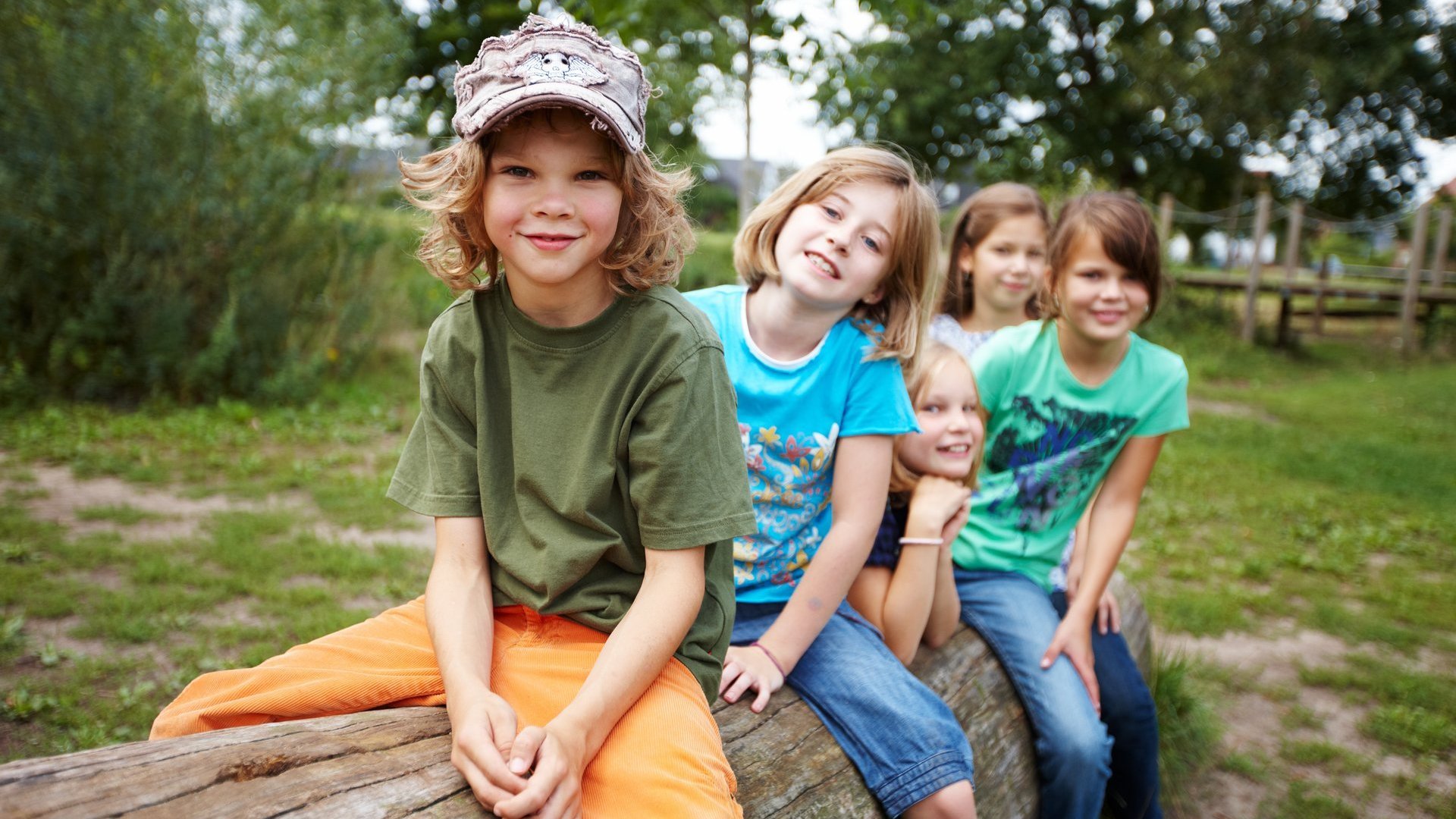 5 Kinder sitzen hintereinander auf einem Baumstamm und lächeln in die Kamera.