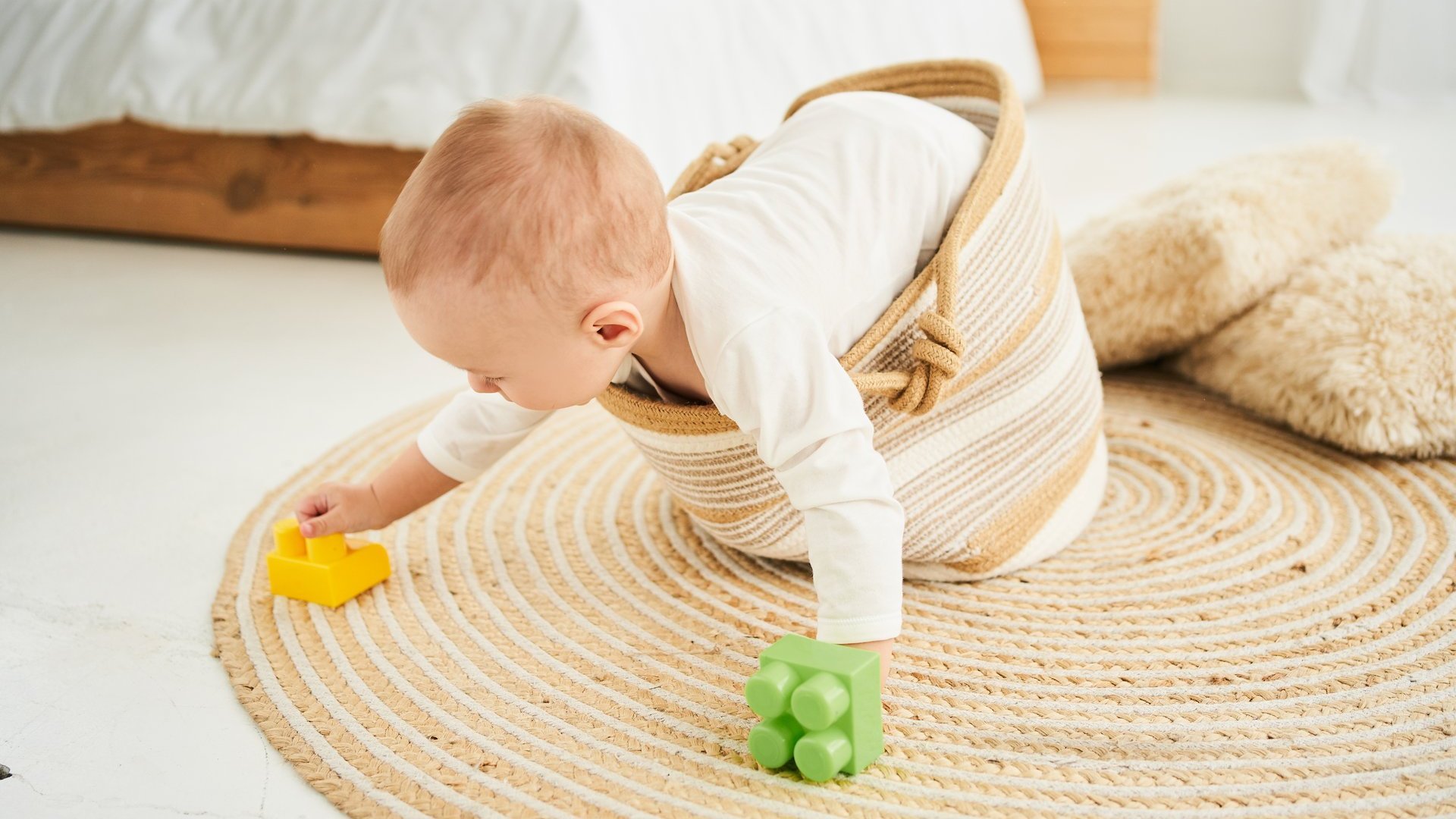 Ein Kleinkind versucht aus einem Korb am Boden rauszuklettern und mit buntem Spielzeug zu spielen.