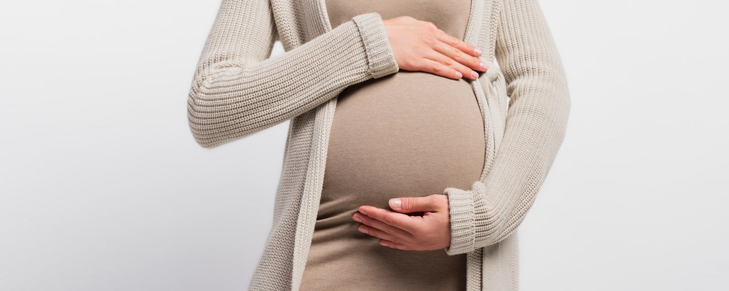 Eine schwangere Frau hält ihren Bauch. Sie trägt ein braunes Kleid und einen beigen Cardigan darüber.