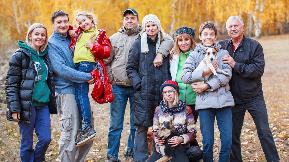 Eine Großfamilie steht mit ihrem Hund im herbstlichen Wald und hält sich im Arm. Alle blicken zur Kamera