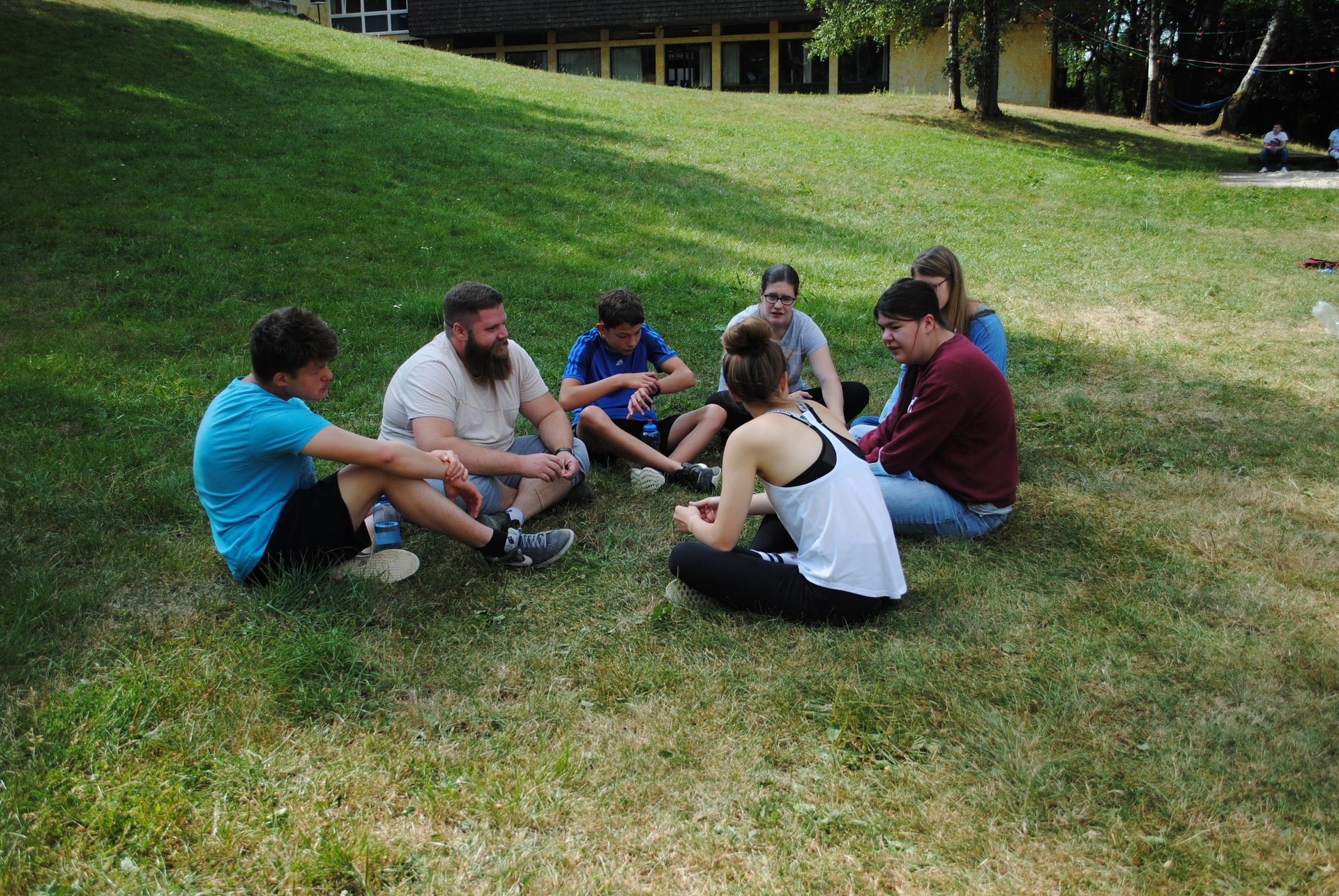Sieben Jugendliche sitzen im Kreis auf einer Wiese und unterhalten sich.