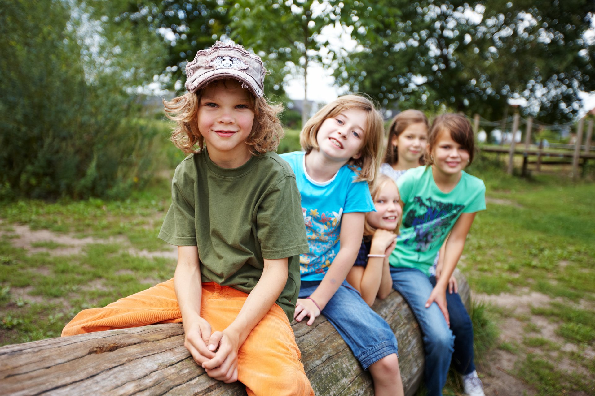 5 Kinder sitzen hintereinander auf einem Baumstamm und lächeln in die Kamera.